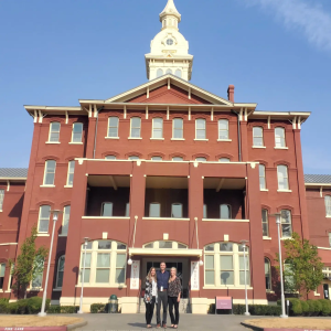 Three people standing in front of brown building that has banners outside its' doorway that says "Museum." front of a